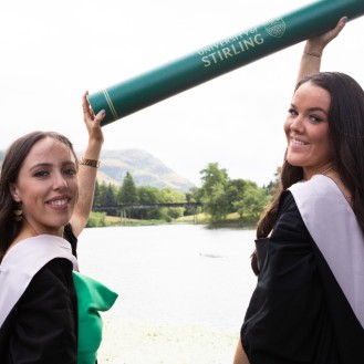 Two female graduates pose by the loch holding a giant scroll prop