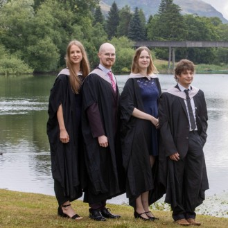 Group of gowned graduates pose with bagpiper at the loch