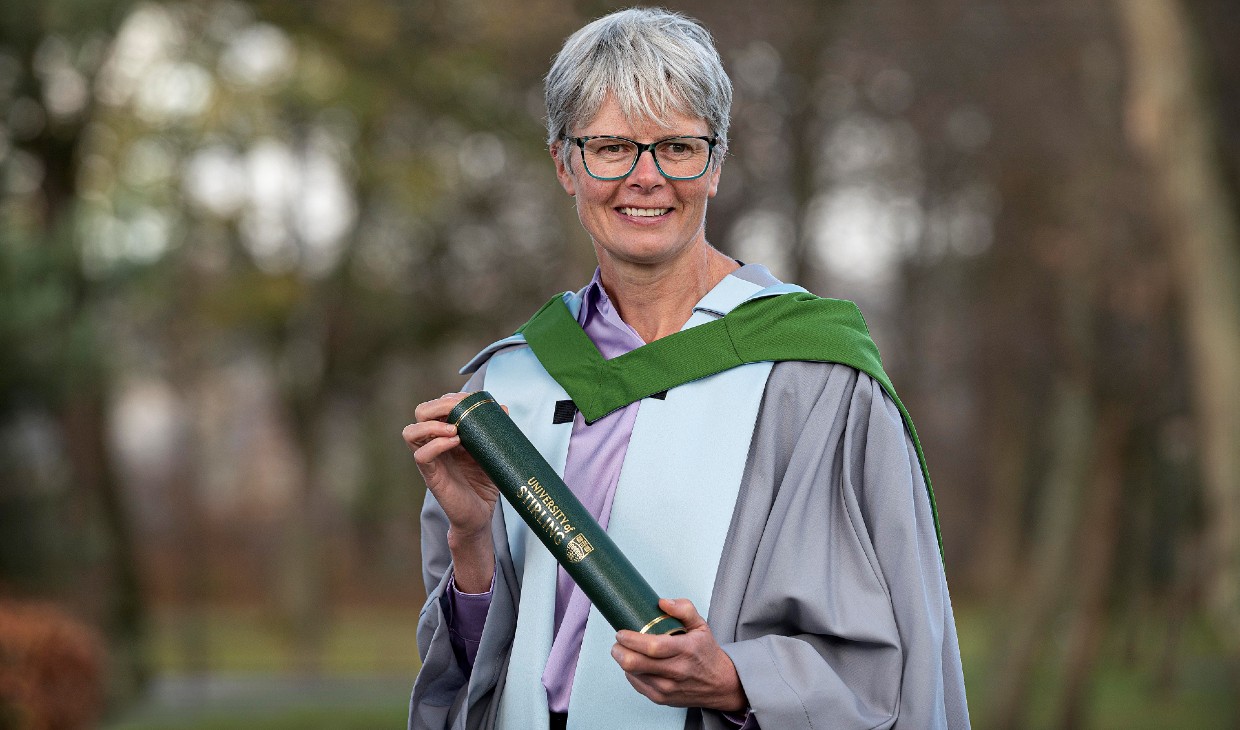 Dr Angela Mudge wears her graduation gown and holds her graduation scroll