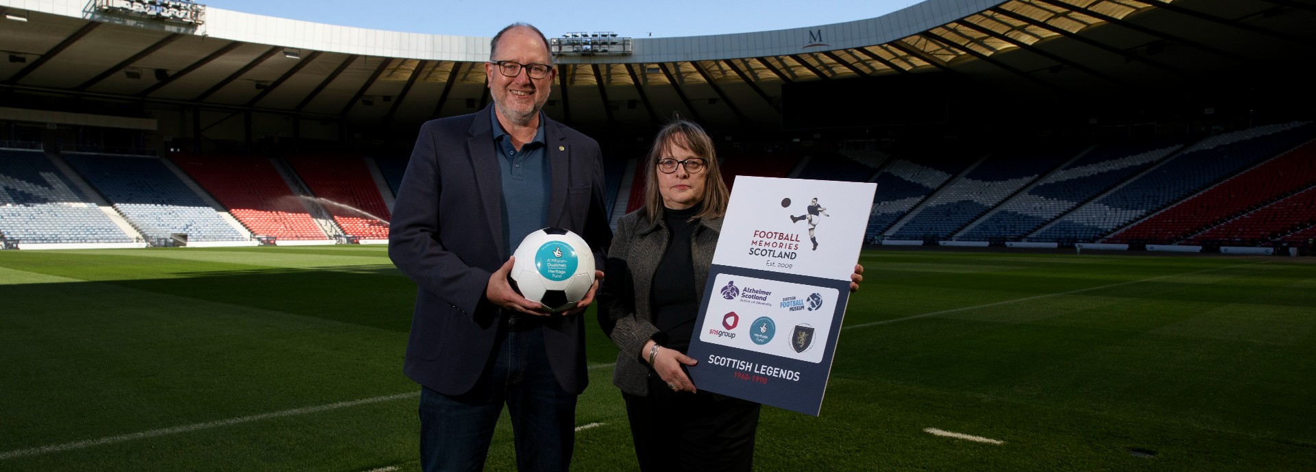 Richard Haynes and Karen Fraser standing on a football pitch with stadium seating in the background. Richard holds a football and Karen holds a large board advertising the 