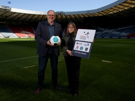 Richard Haynes and Karen Fraser standing on a football pitch with stadium seating in the background. Richard holds a football and Karen holds a large board advertising the 