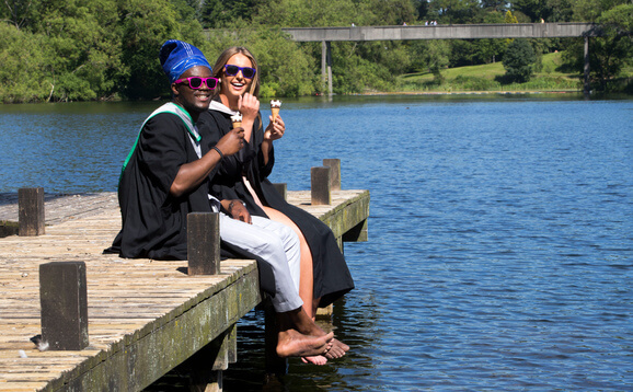 Two graduates eating an ice-cream whilst sitting on a small jetty on a loch