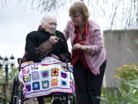 an elderly lady sitting in a wheelchair with a crochet blanket over her lap with her carer outside 