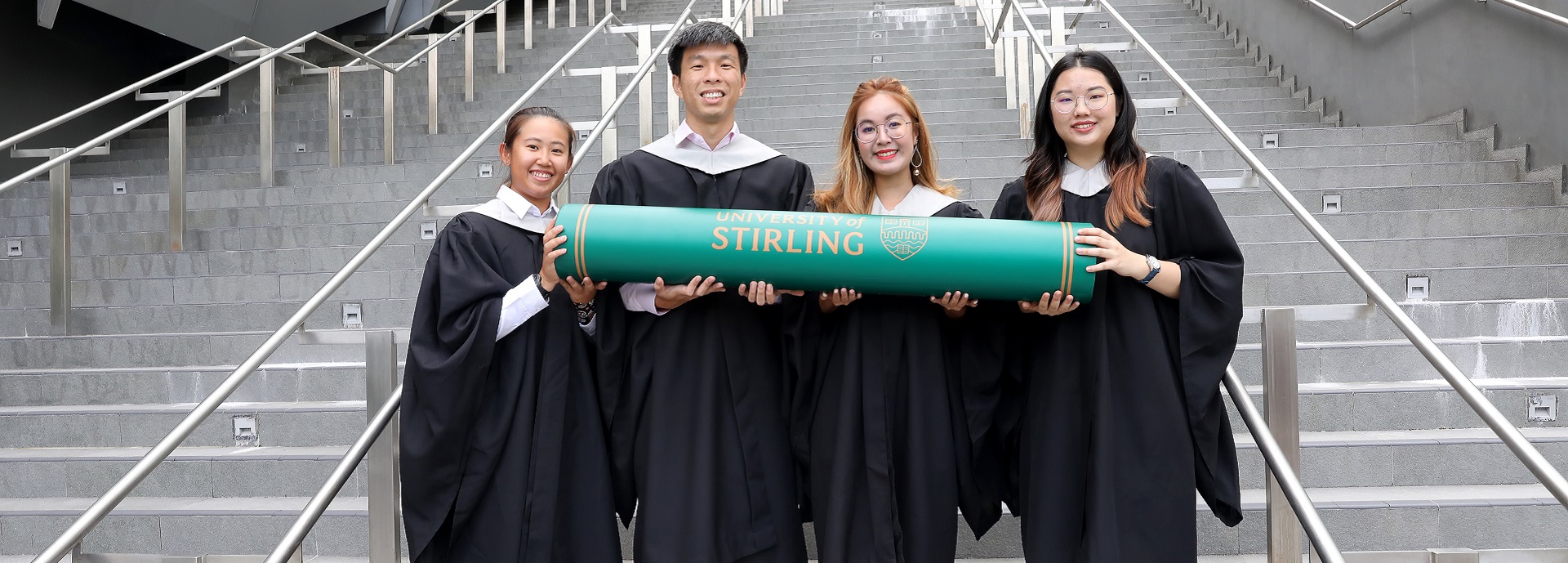 Four Singapore students with graduation robes and giant scroll standing on steps