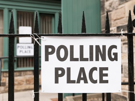 A sign for a polling station tied to a black metal fence with a brick building in the background
