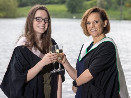 Vicki and Hannah Lawlor wear university graduations gowns and pose next to the loch on Stirling's campus holding champagne glasses