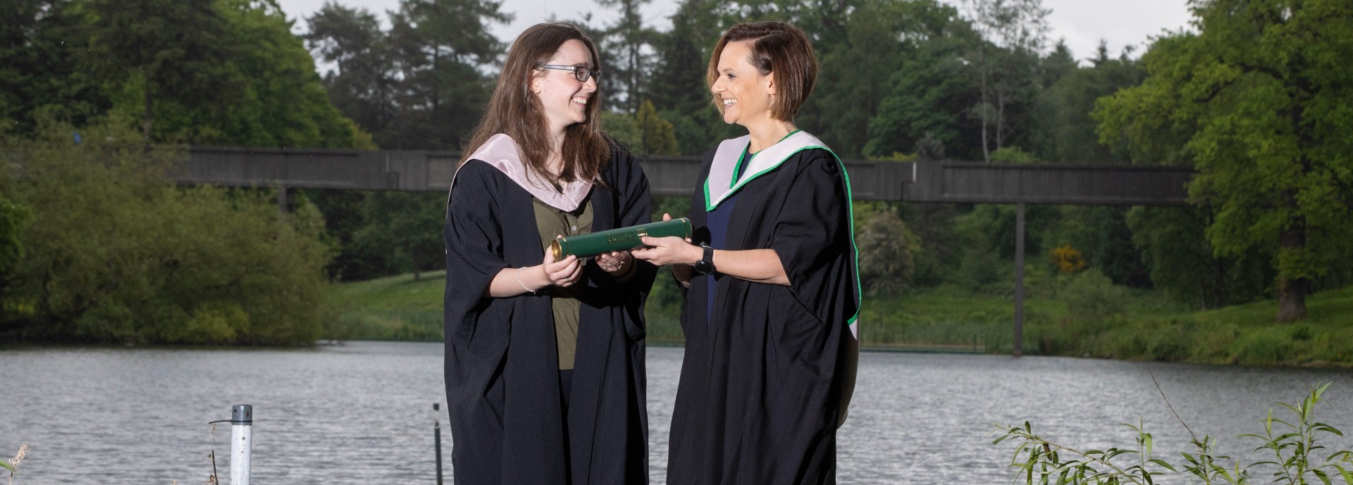 Vicki and Hannah Lawlor wear university graduations gowns and pose next to the loch on Stirling's campus