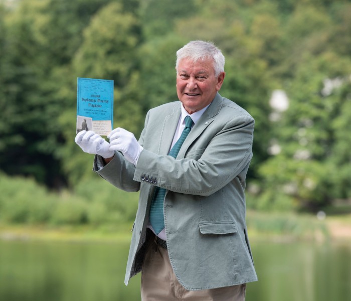 Ron Aitchison poses on campus holding a small blue booklet which is one of the items he has donated to the new archive