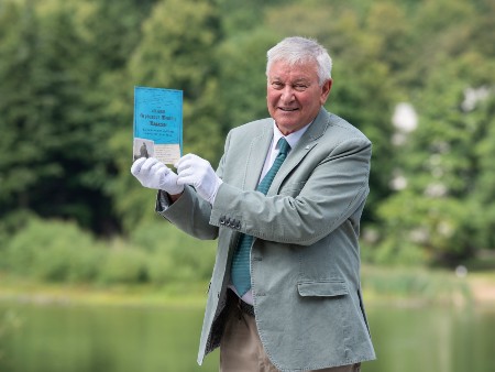 Ron Aitchison poses on campus holding a small blue booklet which is one of the items he has donated to the new archive