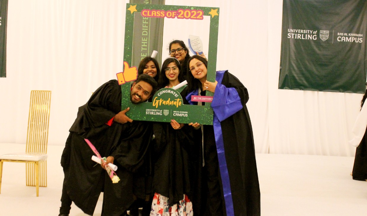 A group of five graduates from Stirling's RAK campus pose using a prop - an oversized green photo frame