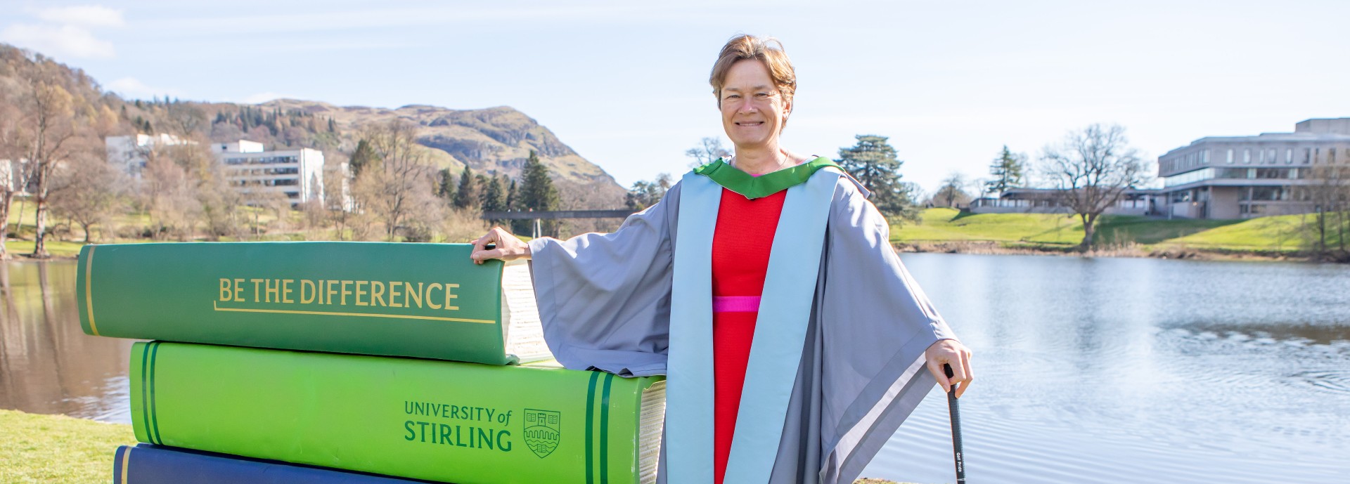 Catriona Mathew wears her graduation gown and poses beside a stack of giant books at the loch on campus