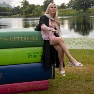 Female graduate perched on large book props by loch