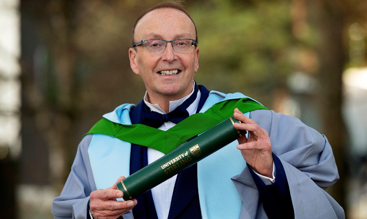 Dr Malcolm Fairweather is pictured outside wearing his graduation robes and holding a scroll