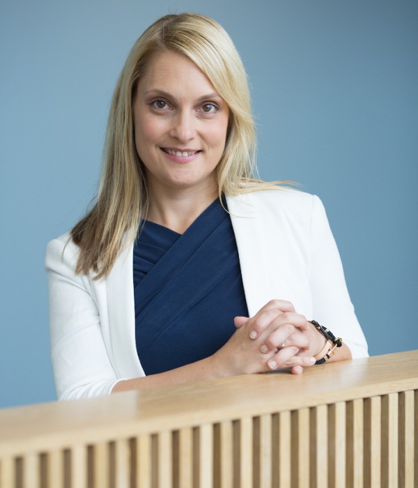 Alenka Jelen leans on a staircase bannister against a blue wall. She has shoulder length blonde hair and is wearing a white suit jacket and navy blue top.