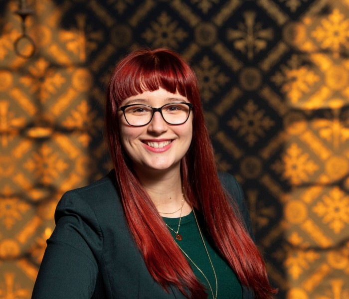 Dr Michaela Hruskova has long red hair with a fringe, black rimmed glasses and is pictured against a patterned background