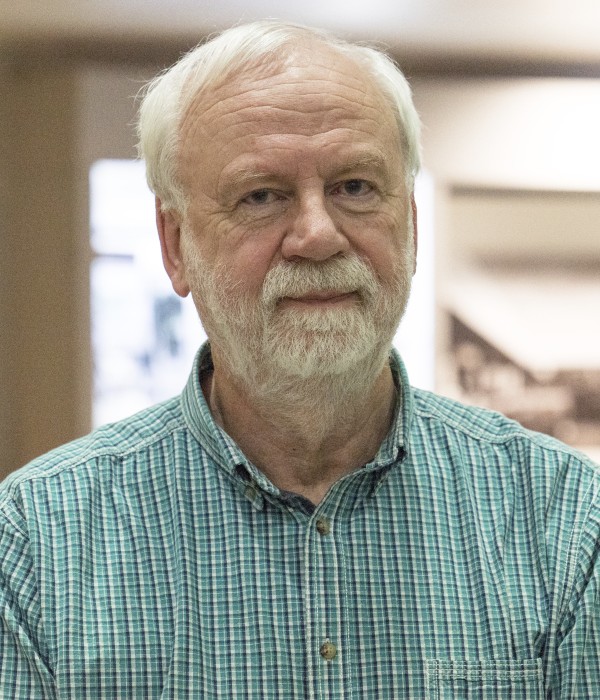 Professor Andrew Watterson is photographed indoors, the background is blurred. Professor Watterson is a white male and wears a green checked shirt, has white hair and a white beard and moustache. He is looking straight to camera.