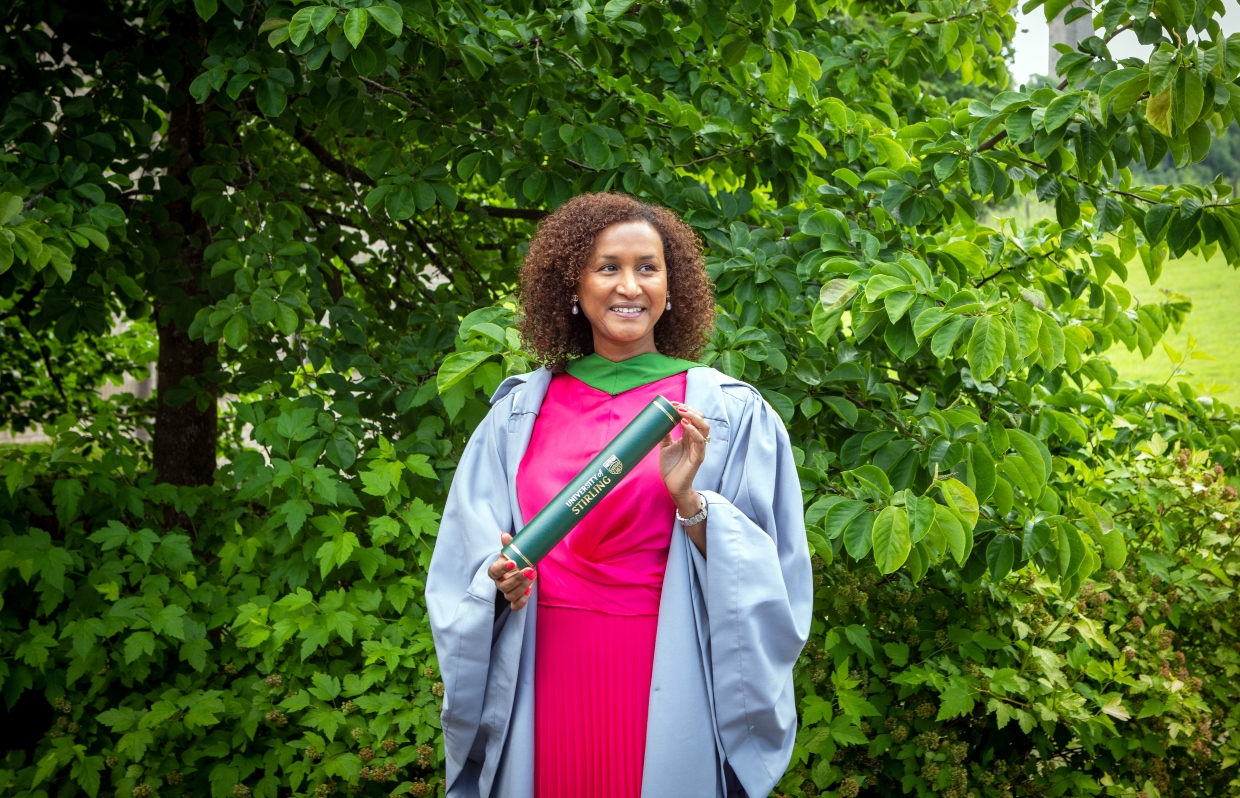 Khadija Coll MBE wears her graduation gown and holds a scroll on campus