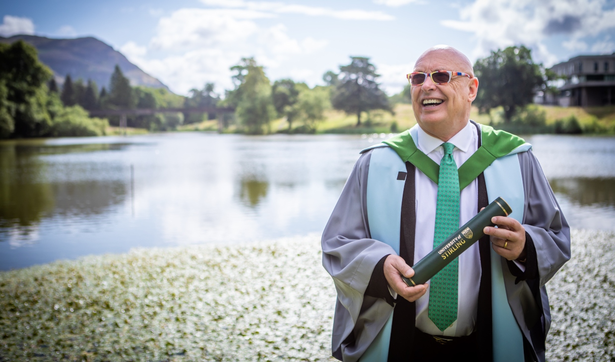 Robert Kilgour wearing graduation robes and holding a graduation scroll photographed on the bank of the campus loch