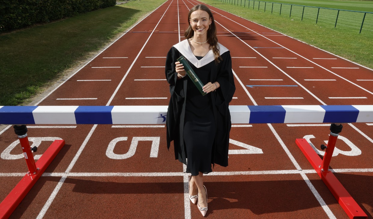 Athlete Sarah Tait poses in her graduation gown on the running track holding her graduation scroll