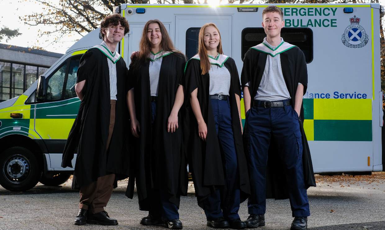 Graduates Drew, Ida, Roisin and Matthew pose in graduation gowns in front of an ambulance