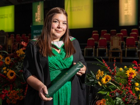 Suzie John poses in front of the stage at her graduation ceremony. She holds a graduation scroll and is wearing a green dress with graduation robes on top. There are flowers to her right hand side on the stage.