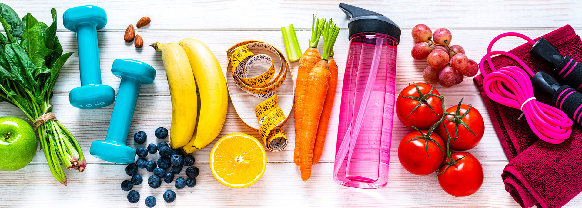 Exercising and healthy eating concept: overhead view of rainbow colored dumbbells, jump rope, water bottle, towel, tape measure and healthy fresh organic vegetables and fruits