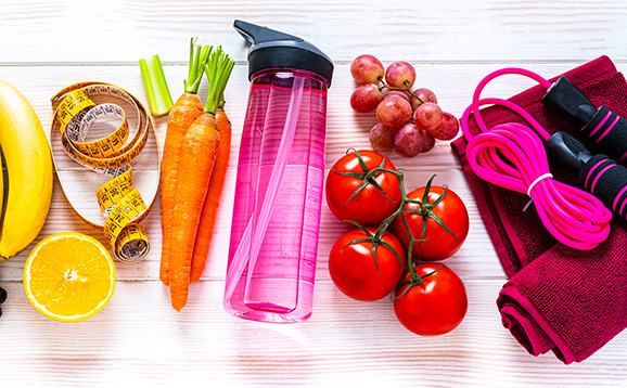 Exercising and healthy eating concept: overhead view of rainbow colored dumbbells, jump rope, water bottle, towel, tape measure and healthy fresh organic vegetables and fruits