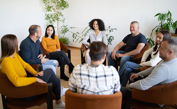 A group of mixed ethnic people sitting in a circle in comfortable chairs holding a support meeting, as they smile and chat with each other